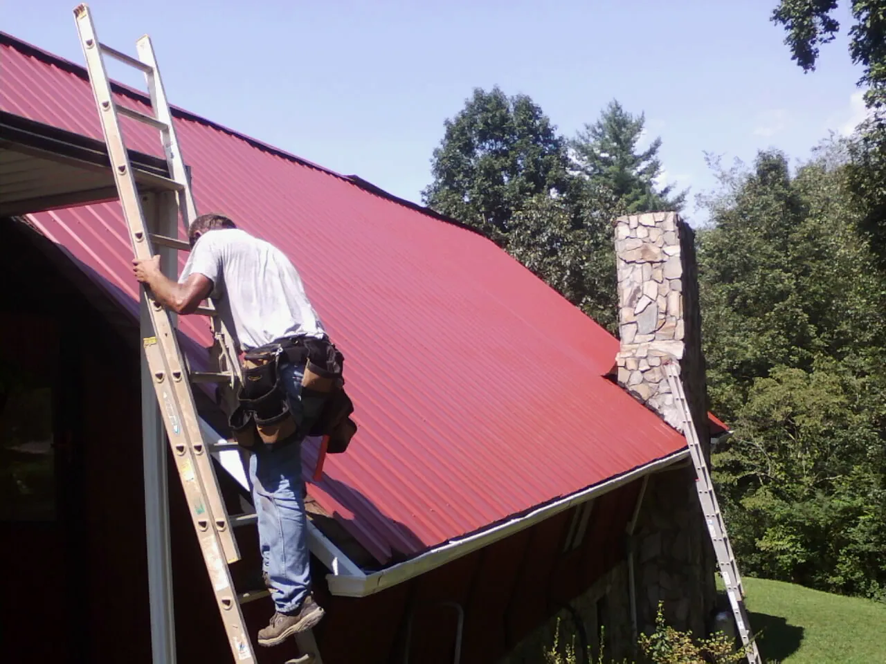 Roofer on a ladder beside a red metal roof and stone chimney — Soffit & Fascia Repair by Roofing Brothers in Lawndale